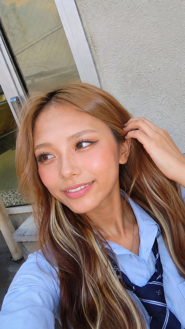 A young woman with long brown and blonde hair, wearing a light blue shirt and a striped tie, smiles while looking to the side in an outdoor setting.