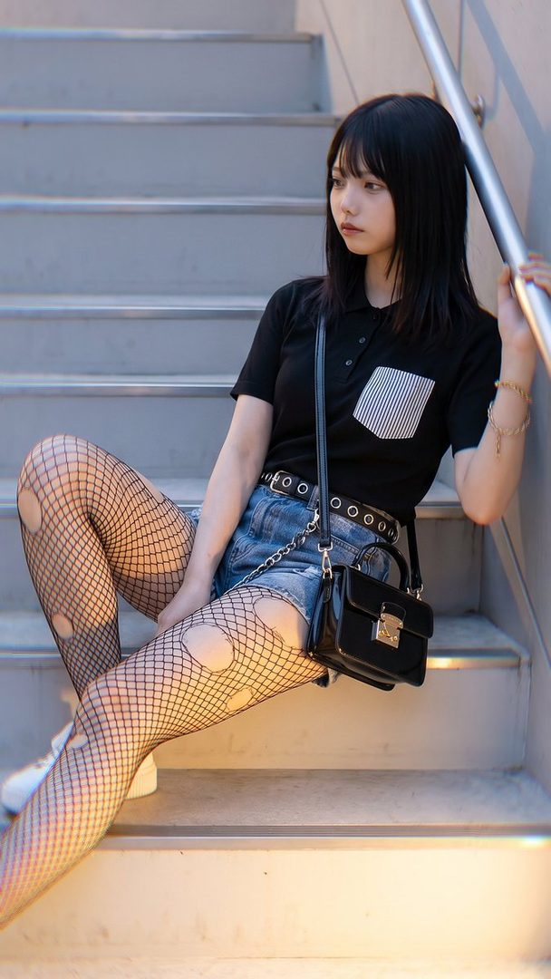 A young woman with black hair and bangs sits on outdoor stairs, wearing a black polo shirt, denim shorts, and ripped fishnet stockings, holding a black handbag.
