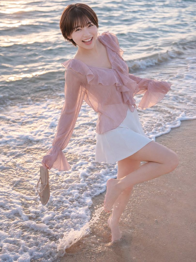 A young woman with short brown hair and a pink ruffled blouse smiles brightly while standing barefoot in the shallow water at a beach during golden hour.