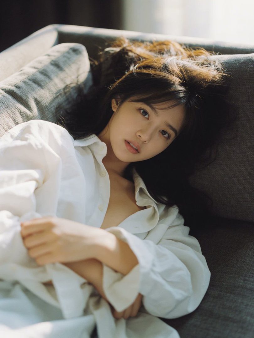 A young woman with long brown hair and a white shirt lies on a sofa, looking dreamily at the camera.
