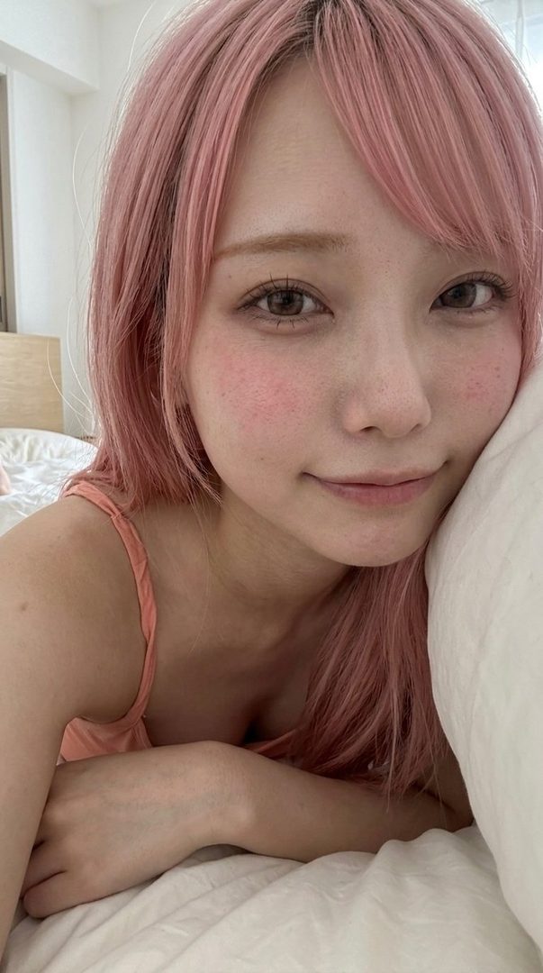 A cute woman with pink hair and bangs smiles softly while lying on a bed, wearing a peach-colored top.