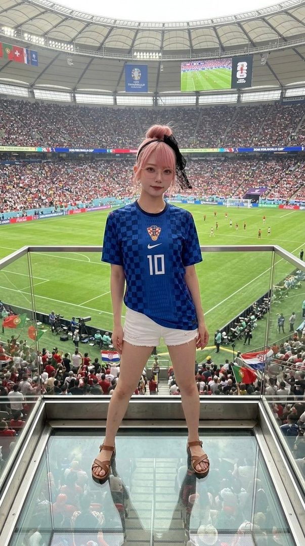 A young woman with pink hair wearing a Croatian soccer jersey and white shorts stands on a glass floor overlooking a stadium during a World Cup match.