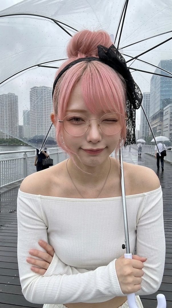 A young woman with pink hair and glasses holds an umbrella on a rainy city street.