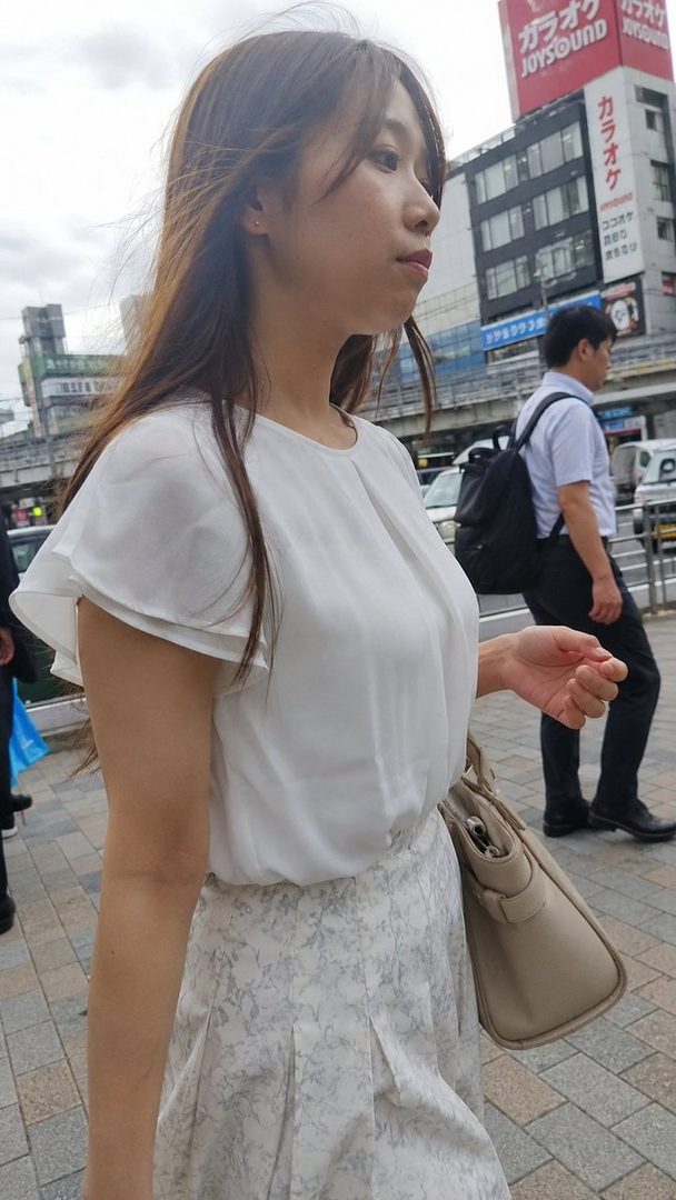A young woman with long brown hair walks on a city street, wearing a white blouse and a patterned skirt, carrying a beige handbag.