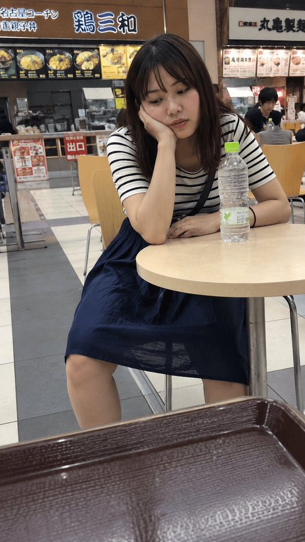 A young woman with long brown hair and bangs, wearing a striped shirt and a dark blue skirt, sits at a table in a food court, resting her chin on her hand with a thoughtful expression.