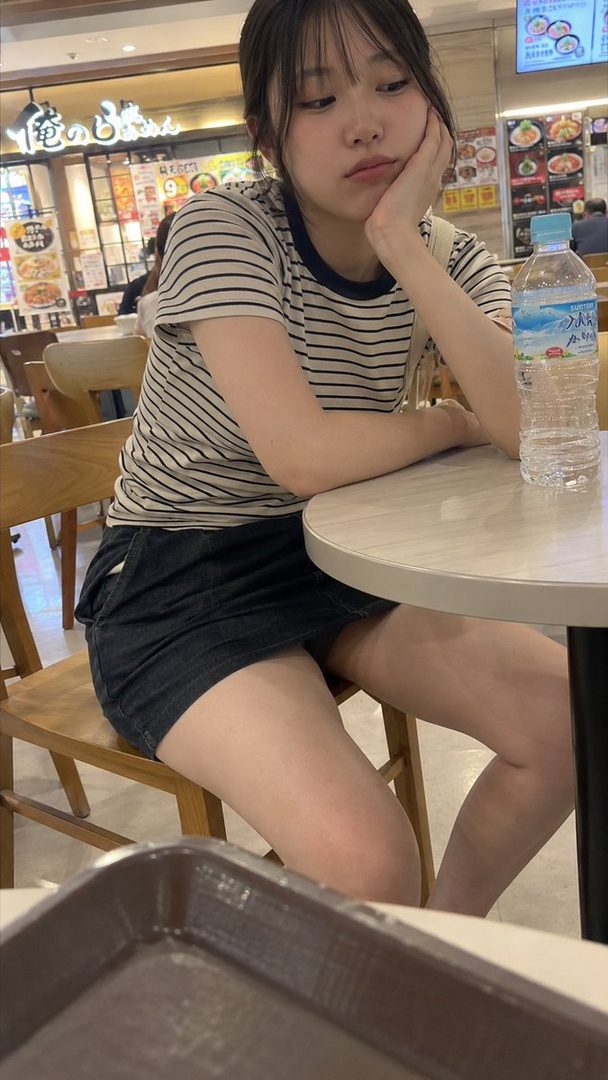 A young East Asian woman with black hair and bangs, wearing a striped t-shirt and denim skirt, sits at a table in a food court, resting her chin on her hand with a thoughtful expression.