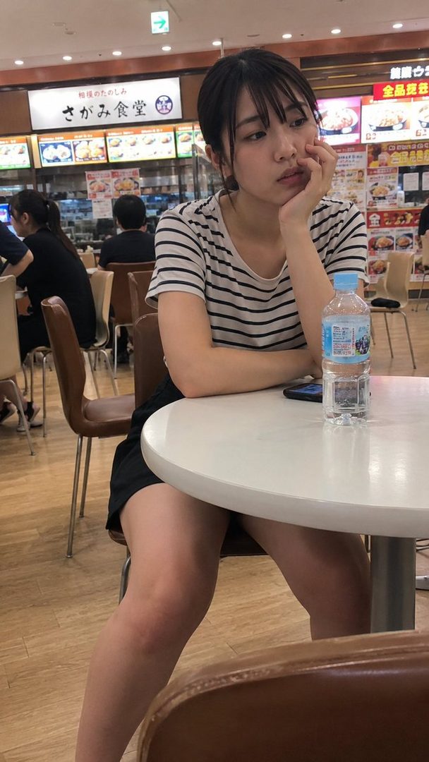 A young woman with black hair and bangs, wearing a striped t-shirt, sits at a table in a food court, looking thoughtful.