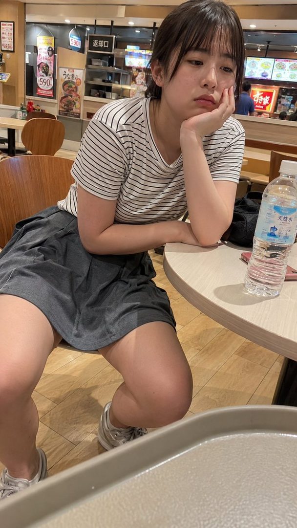 A young woman with short black hair and bangs, wearing a striped t-shirt and a gray skirt, sits at a table in a food court, looking thoughtful.