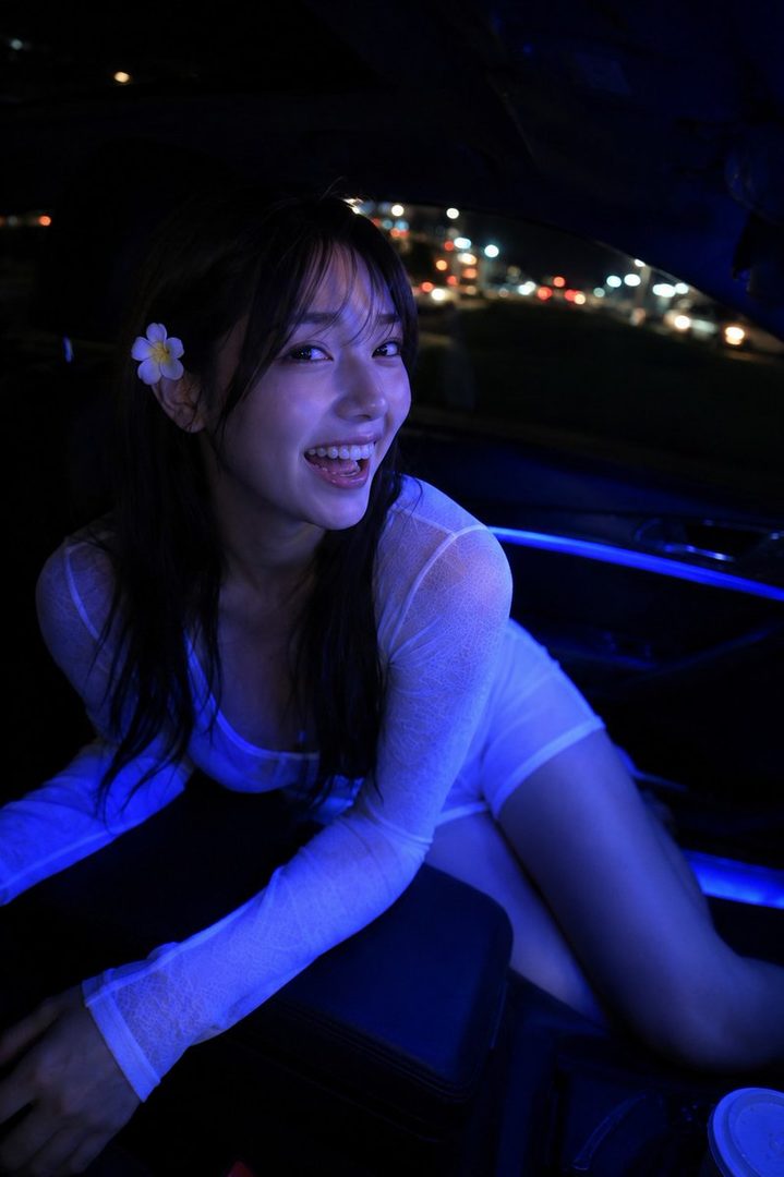 A smiling woman with long dark hair and bangs, wearing a sheer white top, is illuminated by blue light inside a car at night.