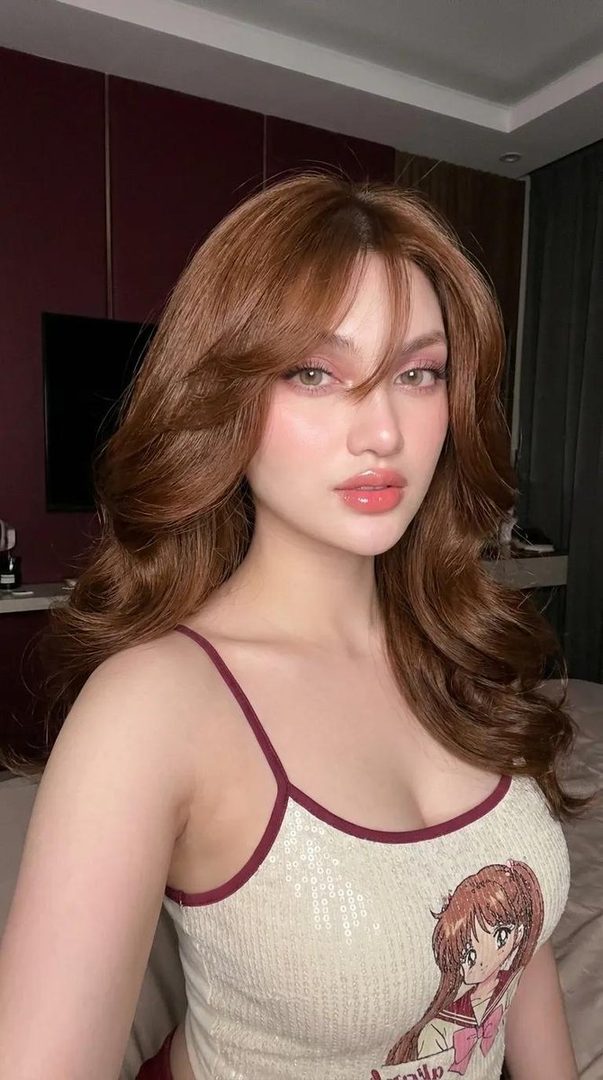 A photorealistic portrait of a woman with long brown hair and bangs, wearing a sequined camisole with an anime character print, posing indoors at night.