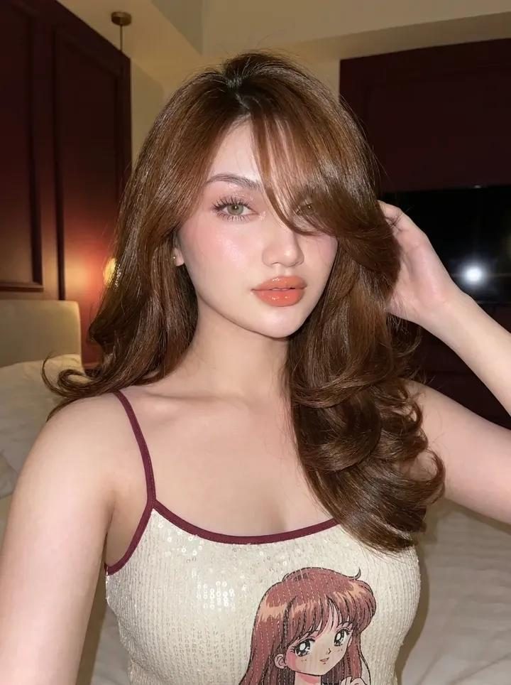A photorealistic portrait of a woman with long brown hair and bangs, wearing a sequined camisole with an anime character print, posing indoors at night.