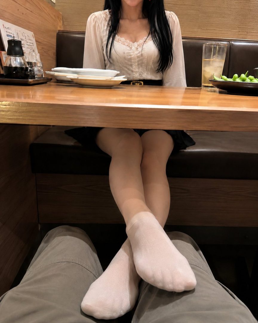A woman with long black hair wearing a white blouse and black skirt sits at a table in a restaurant, with her feet resting on someone's lap.