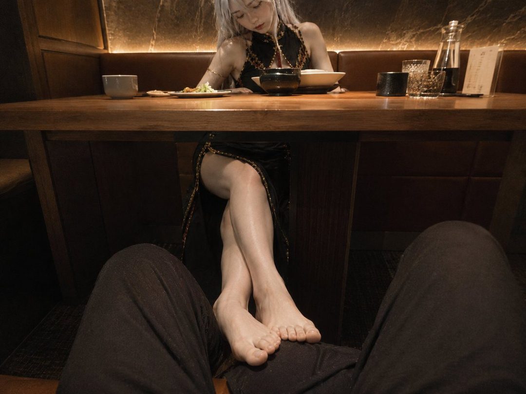 A woman with long white hair wearing a black dress is seated at a table in a dimly lit restaurant, with her bare feet resting on someone's lap under the table.