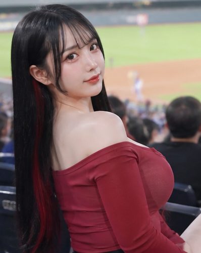 A photorealistic AI-generated image of a young woman with long black hair and red highlights, wearing a red off-the-shoulder top, looking back at the camera from a stadium seat.