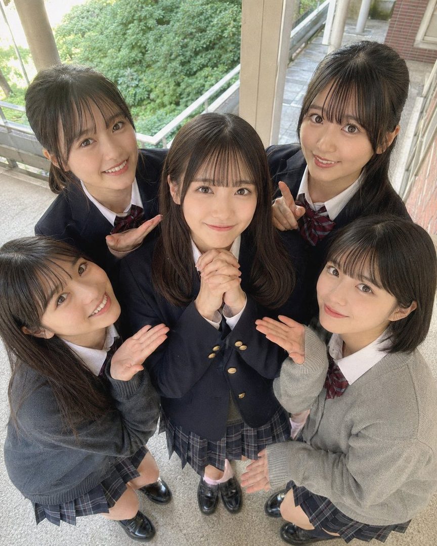 Five young women in Japanese school uniforms pose together, looking up at the camera with smiles.