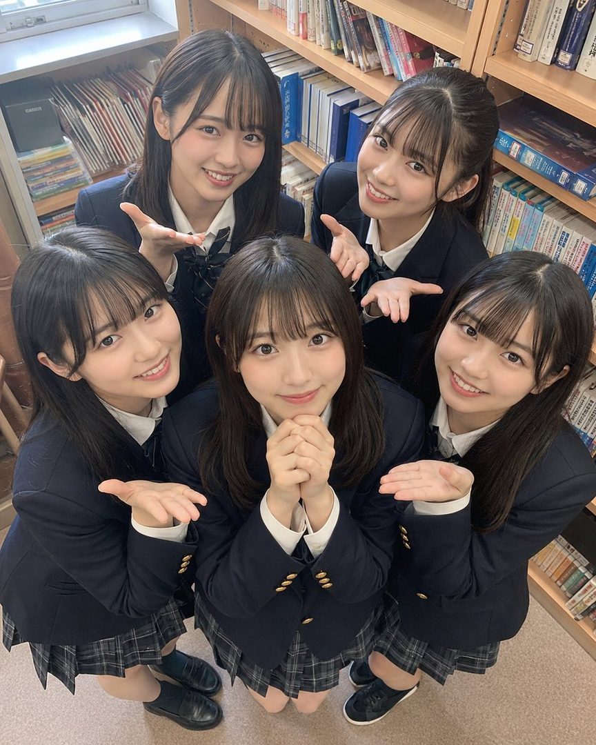 Five young women in school uniforms pose together in a library, looking up at the camera with smiles.