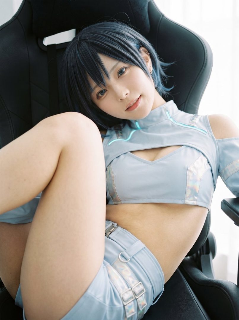 A young woman with blue hair and a light blue crop top and shorts sits in a gaming chair, looking at the camera.
