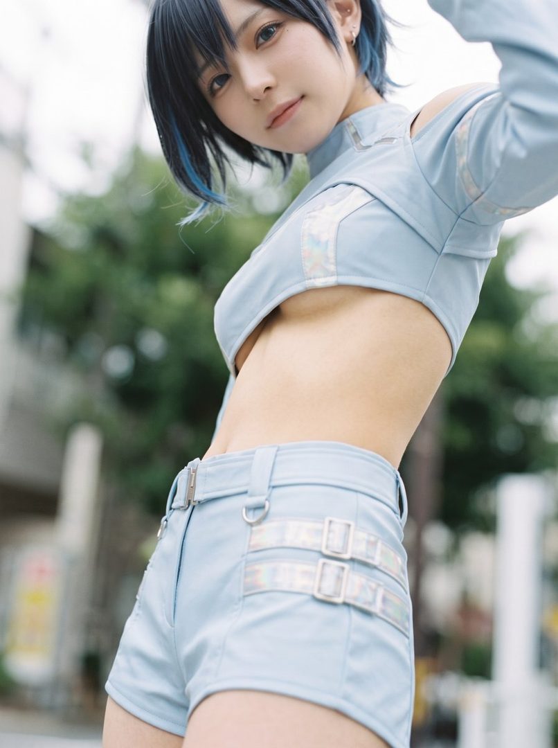 A young woman with short black and blue hair poses outdoors in a light blue crop top and shorts, looking playfully at the camera.