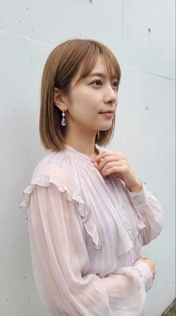 A Japanese woman with short brown hair and bangs wears a light pink ruffled blouse and pearl earrings, looking to the side against a white wall.