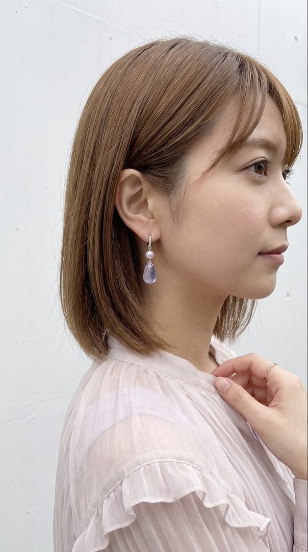 A side profile portrait of a woman with short brown hair and bangs, wearing a light pink ruffled blouse and elegant drop earrings.