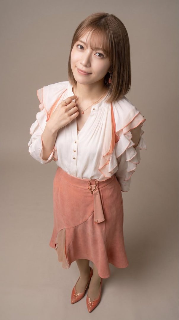 A Japanese woman with short brown hair and bangs smiles gently, wearing a white ruffled blouse and a peach-colored skirt, posing in a studio.