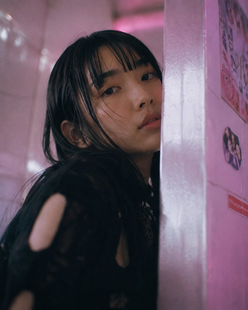 A young woman with black hair and bangs looks directly at the camera, partially obscured by a pink wall.