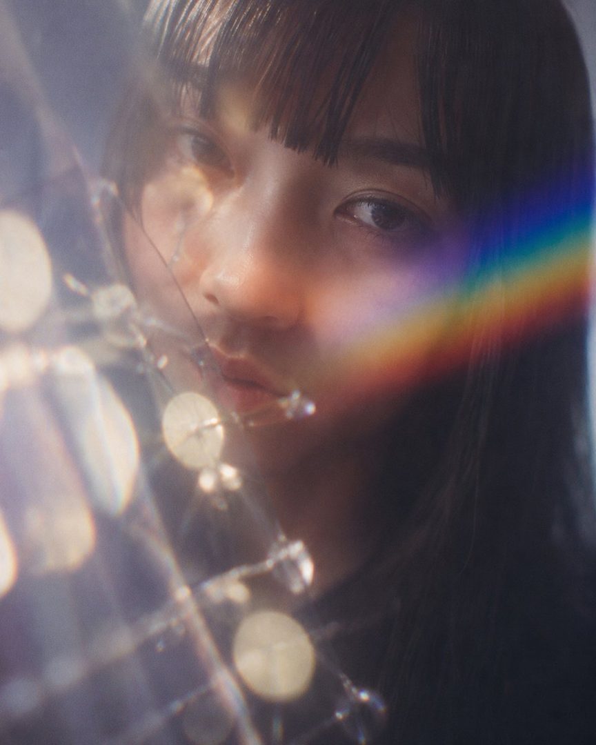 A close-up portrait of a woman with black hair and bangs, looking directly at the camera through a soft, dreamy filter with light flares and a rainbow prism effect.