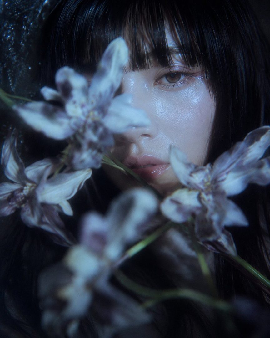 A close-up portrait of a woman with black hair and bangs, partially obscured by delicate purple flowers, with a dreamy and ethereal mood.