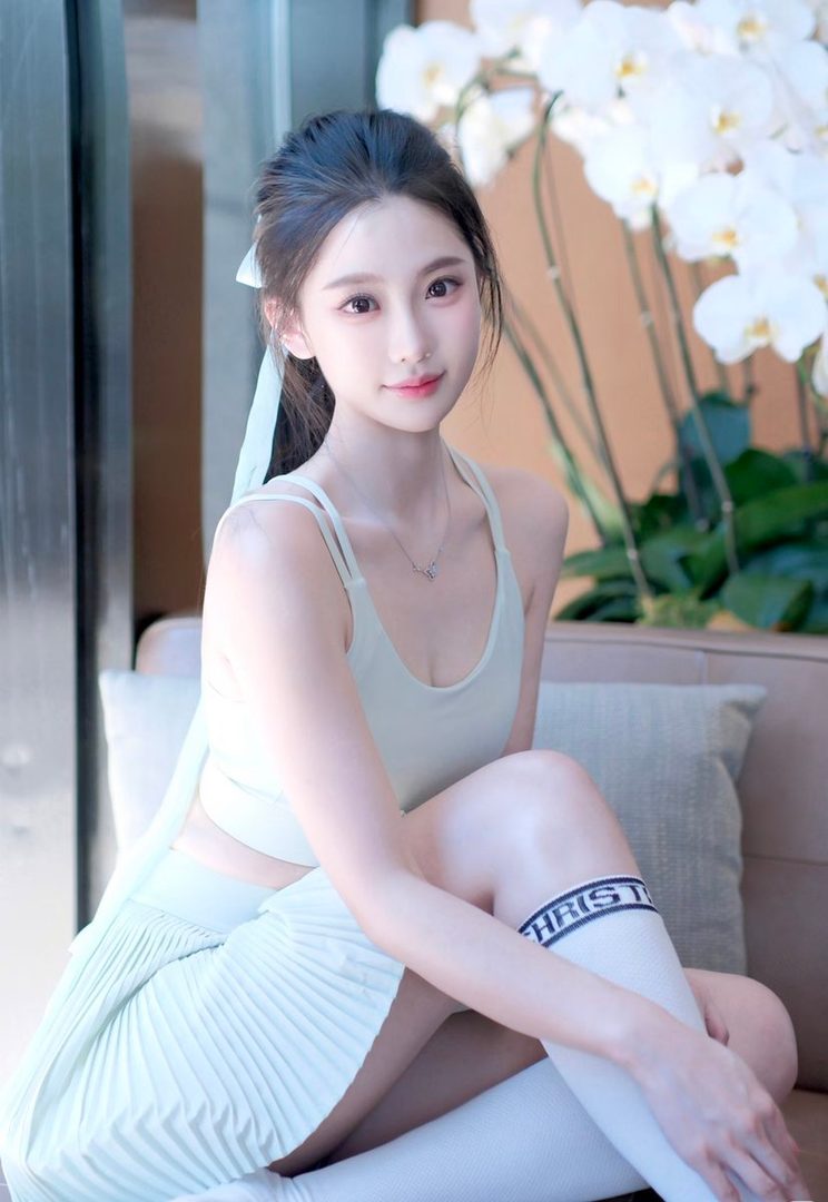 A cute East Asian woman with black hair in a ponytail, wearing a light green sports bra and pleated skirt, sitting indoors with white orchids in the background.