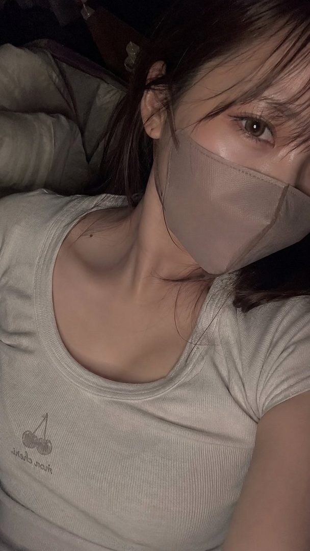 A photorealistic selfie of a young woman with long brown hair, wearing a light-colored ribbed t-shirt and a face mask, looking directly at the camera.