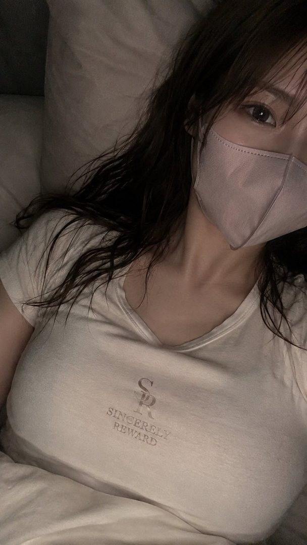 A woman with long brown hair and a face mask takes a selfie while lying down, wearing a white t-shirt.