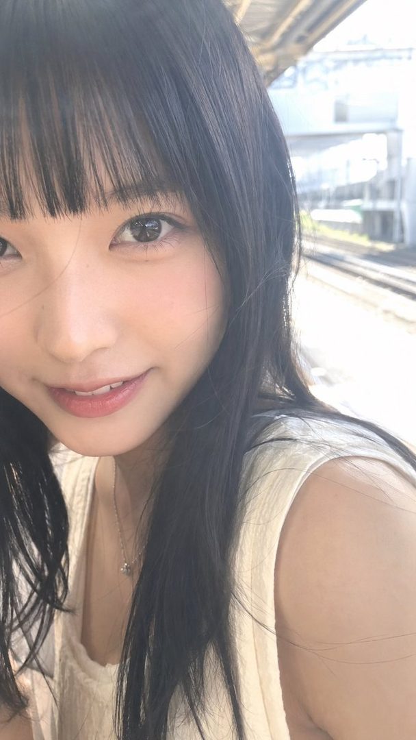A close-up portrait of a young woman with long black hair and bangs, smiling gently at a train station.
