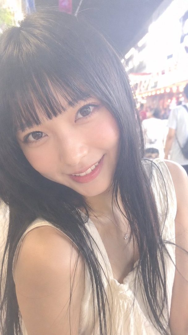 A young woman with long black hair and bangs smiles brightly in a selfie taken outdoors.