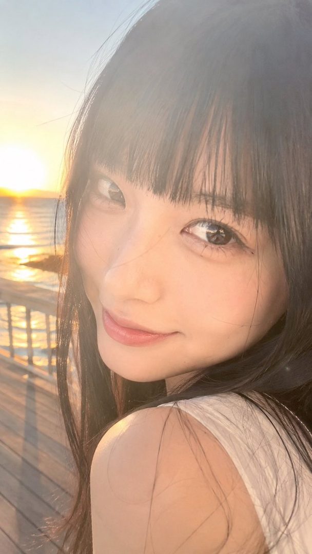 A young woman with long black hair and bangs smiles gently at the camera during golden hour by the sea.