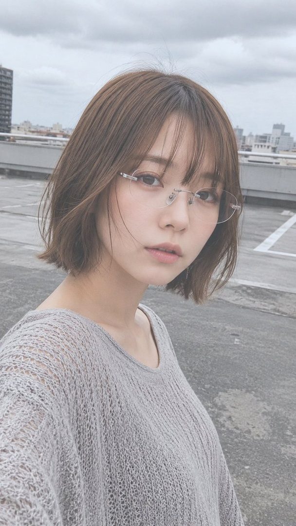A young woman with short brown hair and glasses poses on a rooftop, wearing a light grey knit top.