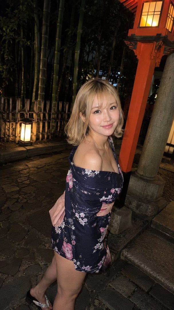A blonde woman in a yukata smiles at the camera at night, with bamboo and a traditional lantern in the background.
