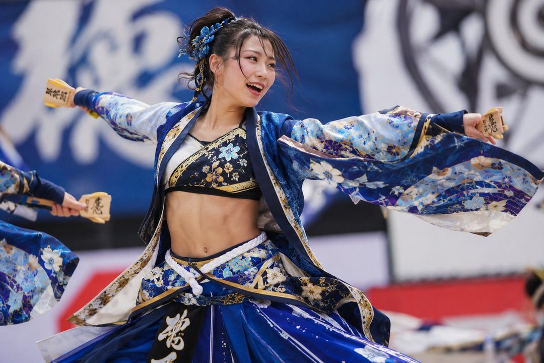 A young woman in a blue and white traditional Japanese-style outfit with floral patterns is captured mid-dance, holding wooden clappers and smiling.