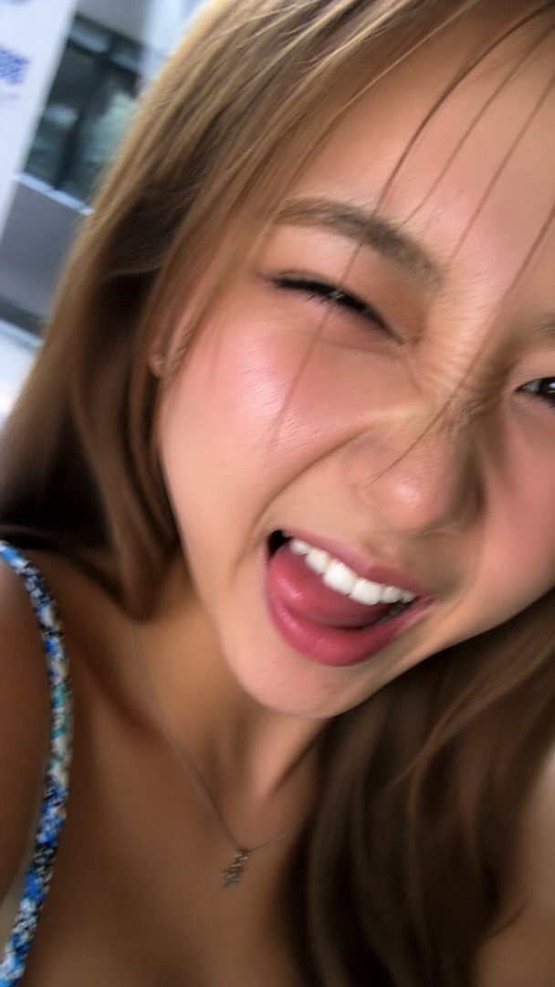 A playful selfie of a young woman with long brown hair, winking and sticking out her tongue, wearing a blue and white patterned top and a delicate necklace.