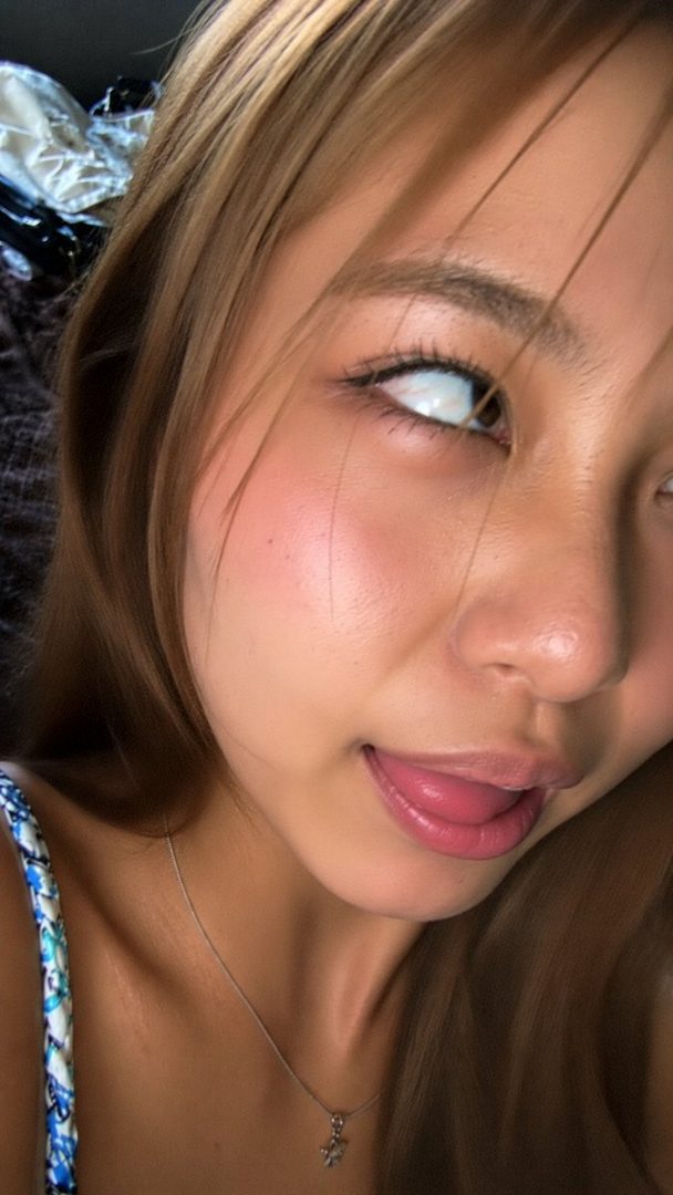 Close-up selfie of a young East Asian woman with long brown hair, sticking her tongue out playfully.