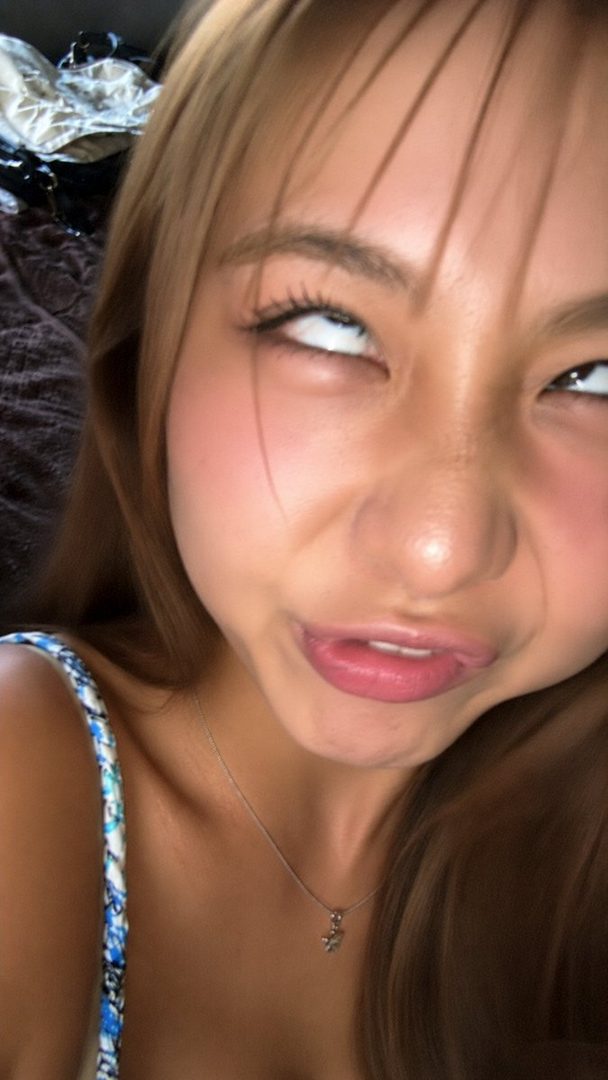 A young woman with long brown hair makes a funny face, rolling her eyes up and sticking out her tongue slightly, wearing a blue and white patterned top and a silver necklace.