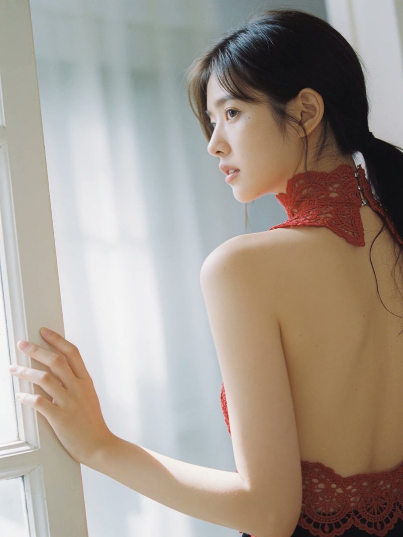 A young woman with black hair in a ponytail, wearing a red lace halter top, looks out a window bathed in soft morning light.