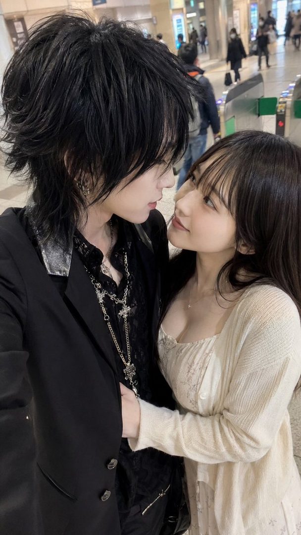 A young couple, a man with dark, styled hair and a woman with long dark hair, looking at each other affectionately in a public place.