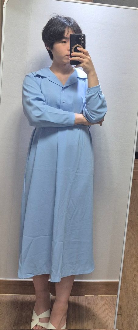 A person with short dark hair takes a mirror selfie, wearing a light blue long-sleeved dress and white sandals.