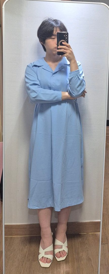 A person with short black hair takes a mirror selfie, wearing a light blue long-sleeved dress and white sandals.