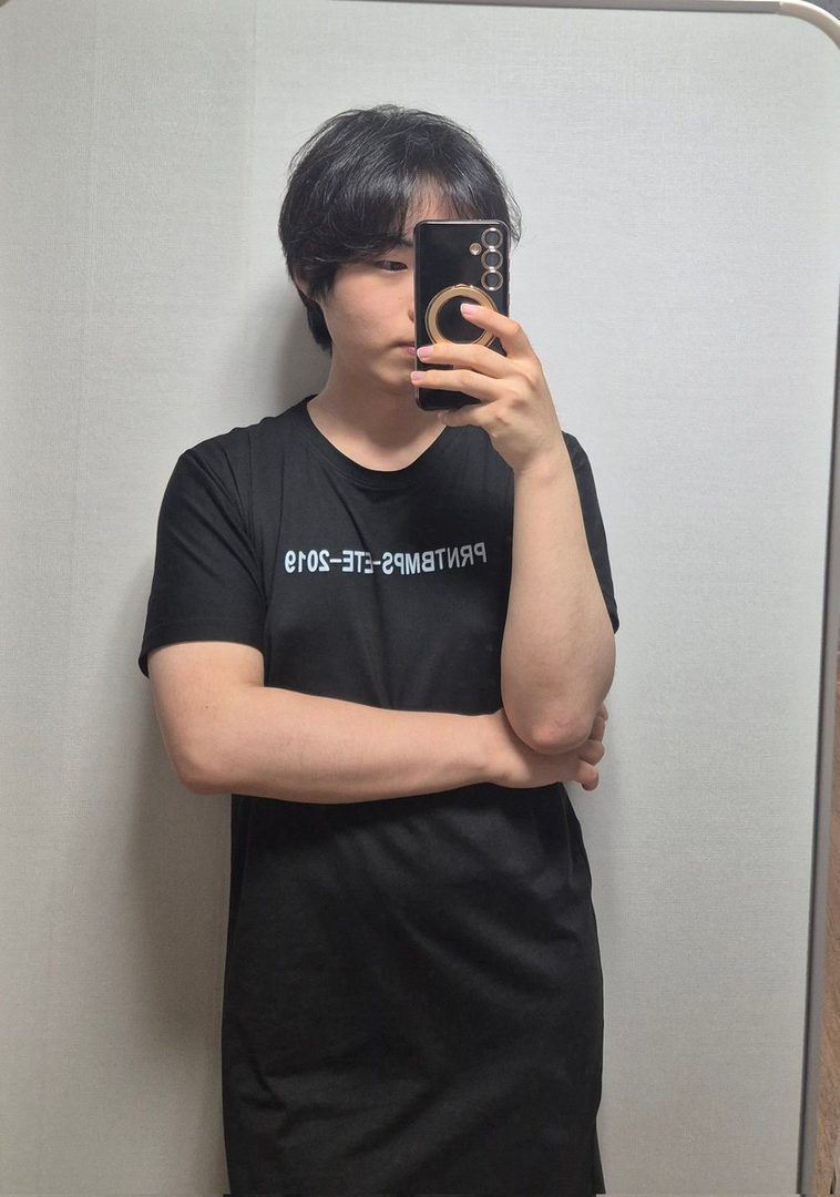 A young East Asian person with short black hair takes a selfie in a black t-shirt, arms crossed.