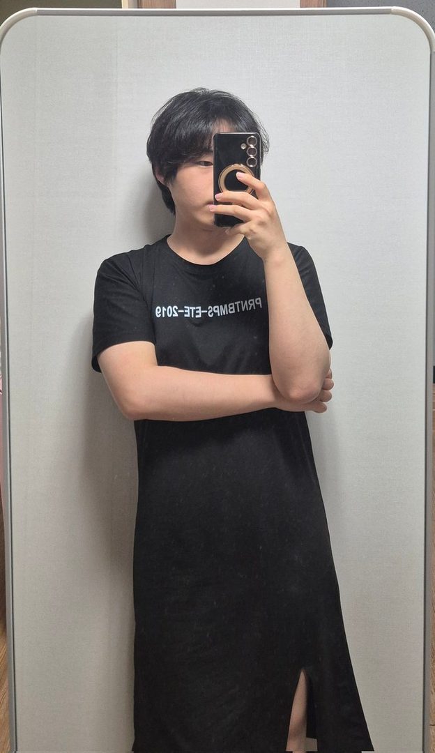 A person with short black hair takes a selfie in a black dress, holding a phone in front of their face.