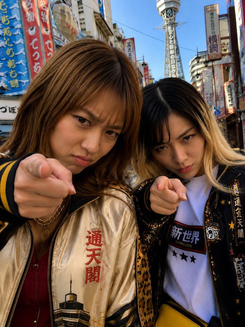 Two young women in embroidered jackets point at the camera on a bustling street in Japan, with a tower in the background.