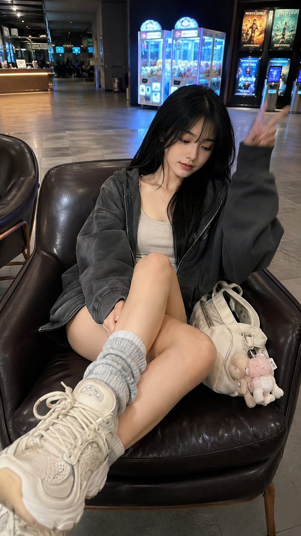 A young East Asian woman with long black hair sits in a leather chair in a cinema lobby, making a peace sign.