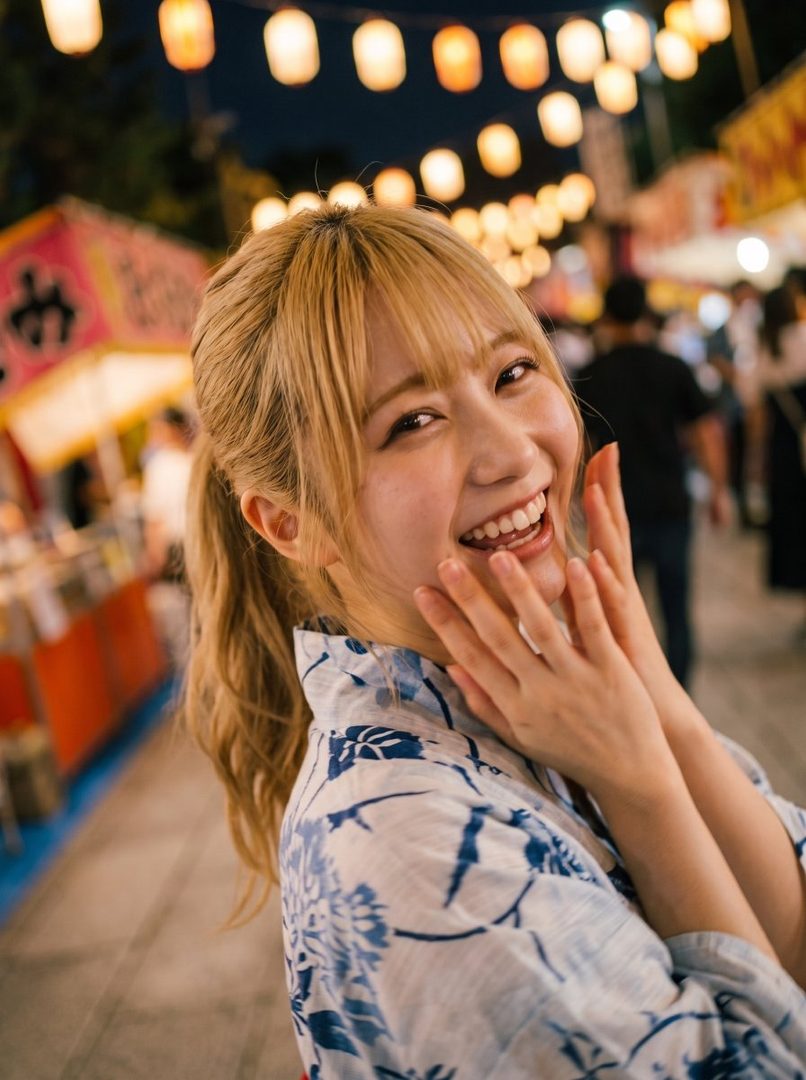 A young woman with blonde hair tied in a ponytail, wearing a blue and white yukata, smiles brightly at a night festival with paper lanterns in the background.
