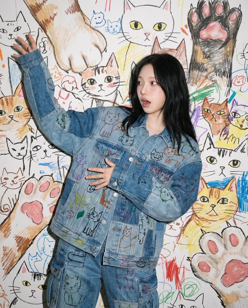 A young woman with long black hair poses in front of a wall covered in colorful cat drawings, wearing a denim jacket and pants adorned with similar cat motifs.
